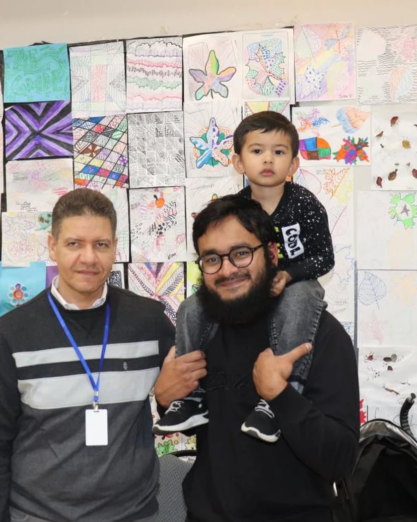 Two parents and a toddler in front of a wall of student artwork